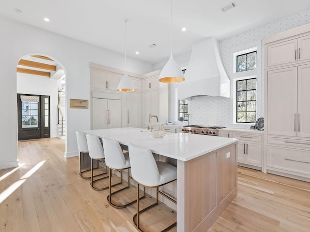 kitchen featuring premium range hood, hanging light fixtures, light hardwood / wood-style flooring, paneled built in fridge, and an island with sink