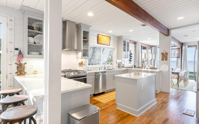 kitchen with appliances with stainless steel finishes, beamed ceiling, wall chimney exhaust hood, kitchen peninsula, and light hardwood / wood-style flooring