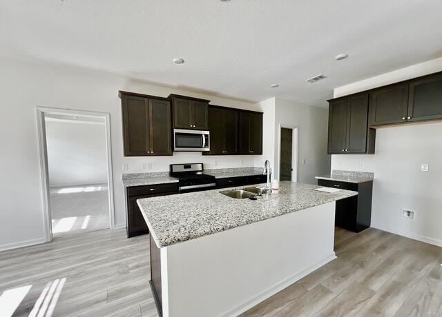 kitchen featuring sink, stainless steel appliances, light hardwood / wood-style floors, light stone countertops, and a center island with sink