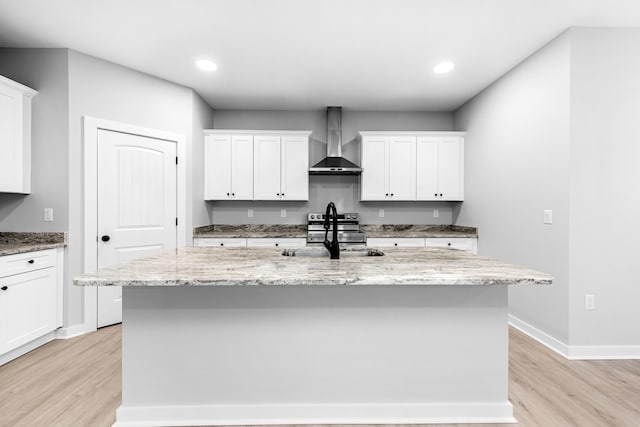 kitchen with wall chimney range hood, sink, light stone counters, an island with sink, and white cabinets