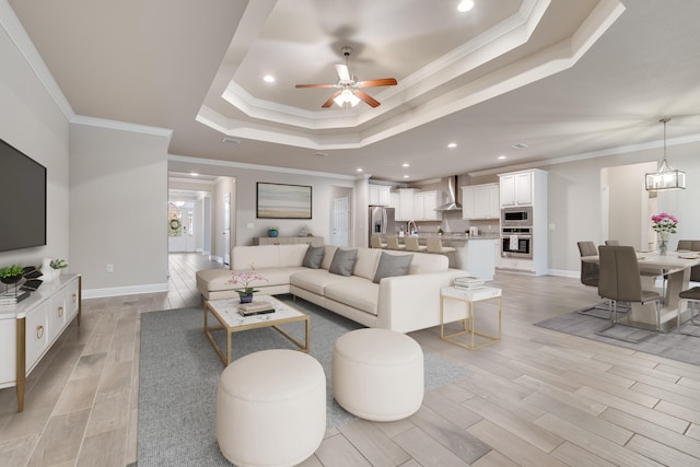 living room with sink, ornamental molding, light hardwood / wood-style floors, ceiling fan with notable chandelier, and a raised ceiling