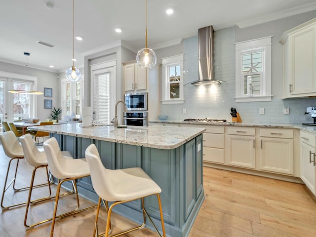 kitchen with light stone counters, a kitchen island with sink, stainless steel appliances, hanging light fixtures, and wall chimney range hood