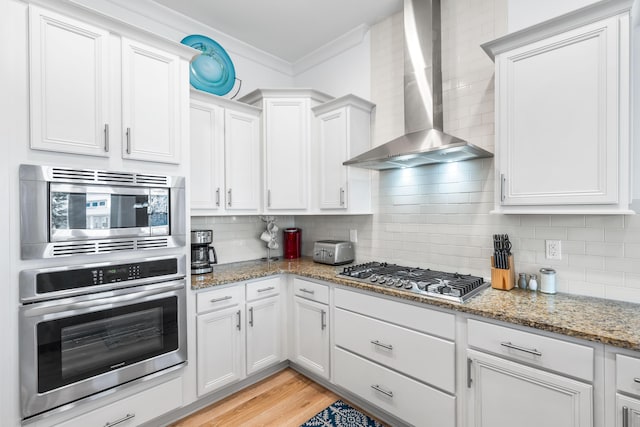 kitchen featuring appliances with stainless steel finishes, white cabinetry, light stone counters, crown molding, and wall chimney range hood