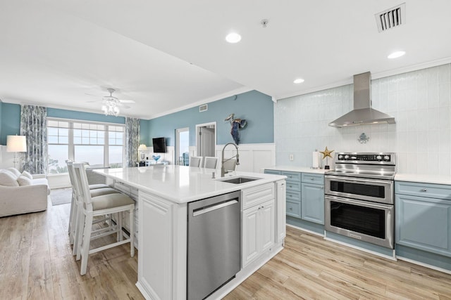 kitchen with white cabinetry, sink, stainless steel appliances, a center island with sink, and wall chimney exhaust hood