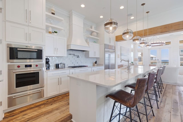 kitchen with pendant lighting, premium range hood, white cabinetry, stainless steel appliances, and an island with sink