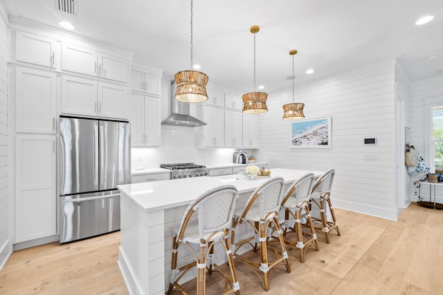 kitchen featuring white cabinetry, stainless steel fridge, an island with sink, pendant lighting, and wall chimney range hood