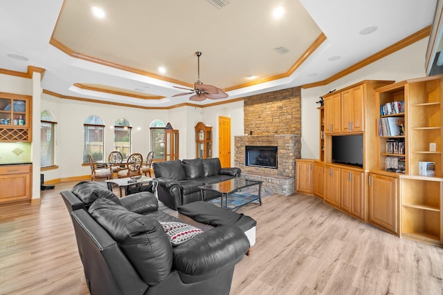 living room featuring a stone fireplace, light wood-type flooring, ornamental molding, a raised ceiling, and ceiling fan