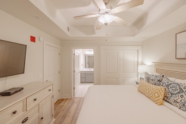 bedroom featuring ceiling fan, ensuite bath, light hardwood / wood-style floors, and a tray ceiling