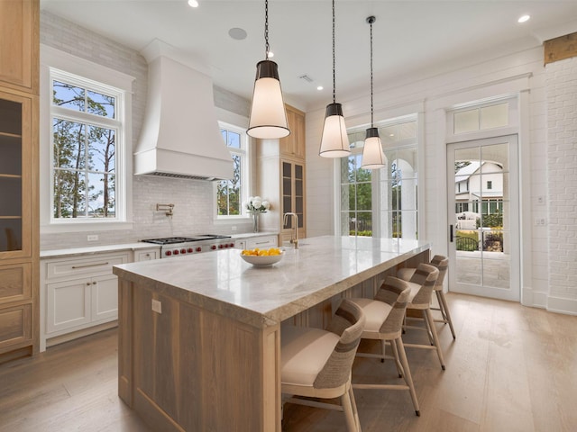 kitchen featuring a kitchen island with sink, light hardwood / wood-style floors, custom range hood, white cabinets, and stainless steel gas cooktop