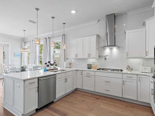 kitchen with appliances with stainless steel finishes, white cabinetry, sink, hanging light fixtures, and wall chimney range hood