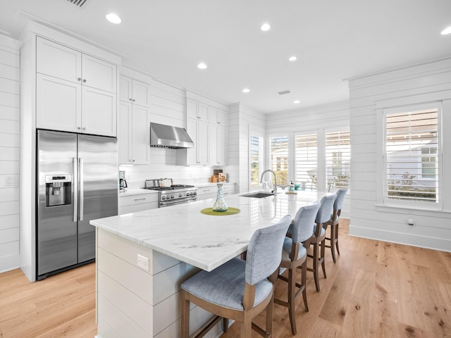 kitchen featuring white cabinets, a kitchen bar, a kitchen island with sink, stainless steel appliances, and wall chimney exhaust hood