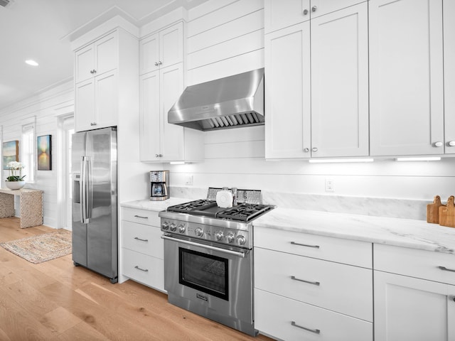 kitchen with wall chimney exhaust hood, white cabinetry, light stone counters, appliances with stainless steel finishes, and light hardwood / wood-style floors