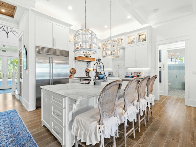 kitchen featuring built in refrigerator, white cabinets, hanging light fixtures, a center island with sink, and glass insert cabinets