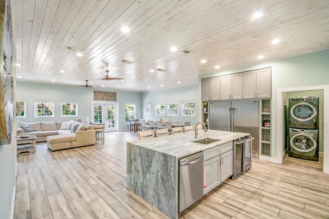 kitchen featuring dishwasher, light stone countertops, a kitchen island with sink, stacked washing maching and dryer, and a sink