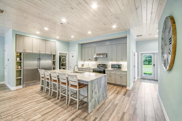 kitchen with stainless steel appliances, stacked washer and dryer, gray cabinetry, a kitchen island with sink, and a sink