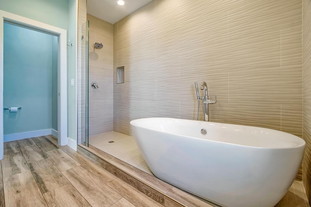 full bathroom featuring baseboards, wood finished floors, a freestanding tub, a shower stall, and tile walls