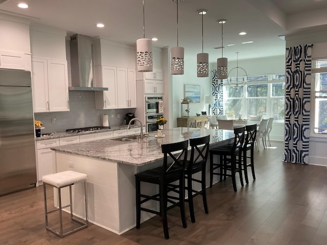 kitchen featuring wall chimney range hood, stainless steel appliances, an island with sink, and white cabinets