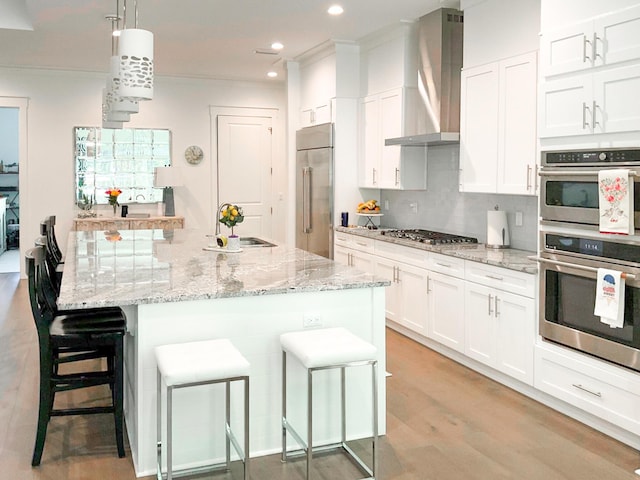 kitchen with hanging light fixtures, wall chimney range hood, a kitchen breakfast bar, stainless steel appliances, and white cabinets