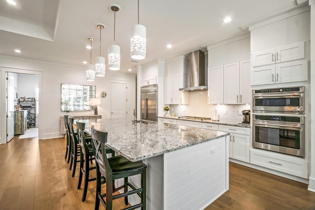 kitchen featuring white cabinetry, hanging light fixtures, a kitchen island with sink, stainless steel appliances, and wall chimney range hood