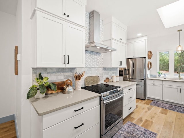 kitchen featuring wall chimney exhaust hood, a skylight, hanging light fixtures, appliances with stainless steel finishes, and white cabinets