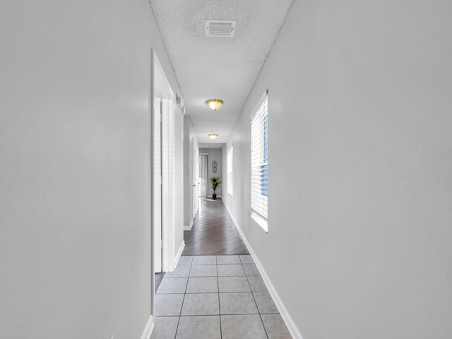 hallway with visible vents, a textured ceiling, baseboards, and light tile patterned flooring