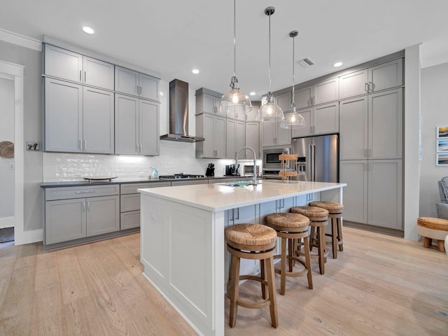 kitchen with wall chimney exhaust hood, sink, appliances with stainless steel finishes, pendant lighting, and a kitchen island with sink