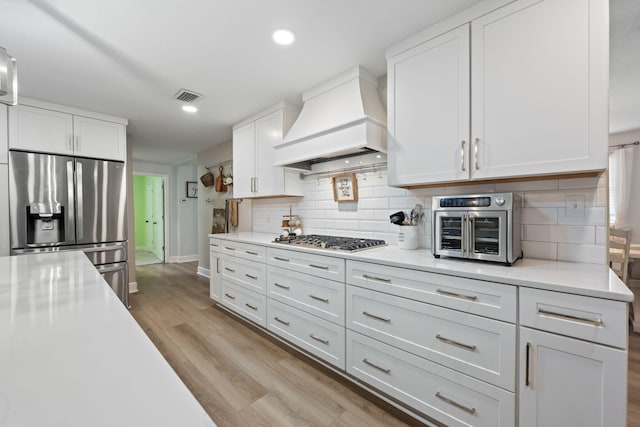 kitchen with stainless steel appliances, premium range hood, visible vents, light wood-style floors, and light countertops