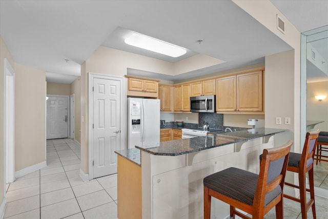 kitchen with light brown cabinetry, a kitchen breakfast bar, dark stone counters, kitchen peninsula, and white appliances