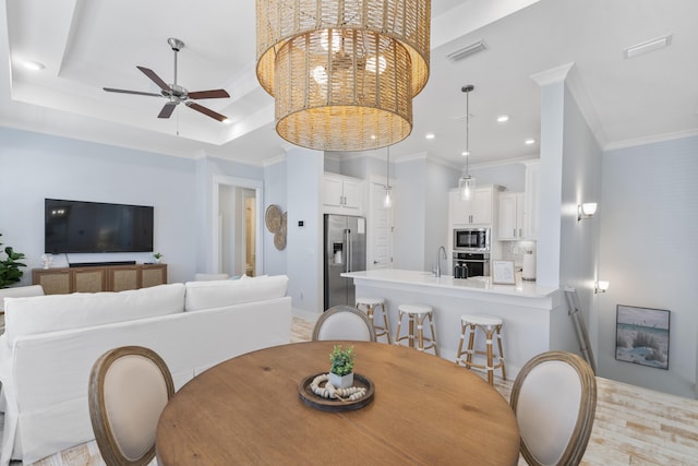 dining room with sink, crown molding, light hardwood / wood-style floors, ceiling fan with notable chandelier, and a raised ceiling