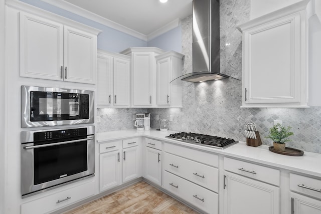 kitchen featuring white cabinetry, tasteful backsplash, ornamental molding, appliances with stainless steel finishes, and wall chimney range hood