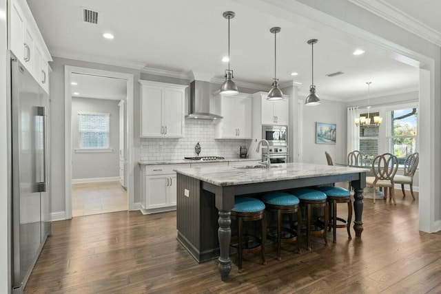 kitchen with wall chimney range hood, appliances with stainless steel finishes, white cabinetry, a center island with sink, and decorative light fixtures