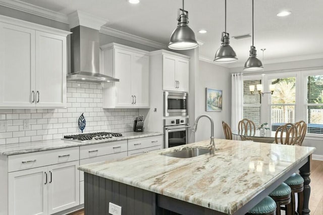 kitchen with appliances with stainless steel finishes, white cabinetry, hanging light fixtures, a center island with sink, and wall chimney exhaust hood