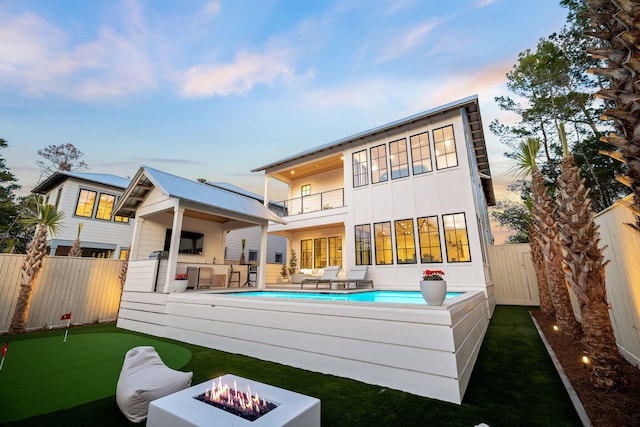 back house at dusk featuring a fenced in pool, a patio area, a balcony, and an outdoor fire pit