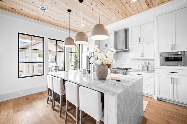 kitchen with a kitchen island with sink, wall chimney range hood, white cabinets, and light stone counters