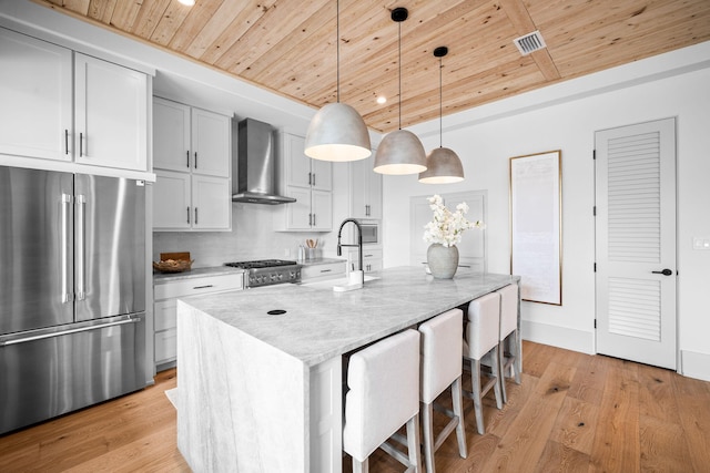 kitchen with pendant lighting, a kitchen island with sink, light stone counters, high end fridge, and wall chimney exhaust hood