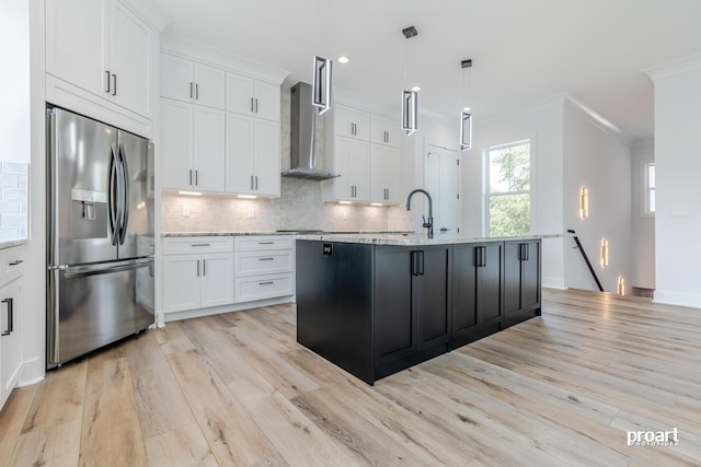 kitchen featuring white cabinetry, stainless steel fridge with ice dispenser, hanging light fixtures, an island with sink, and wall chimney range hood