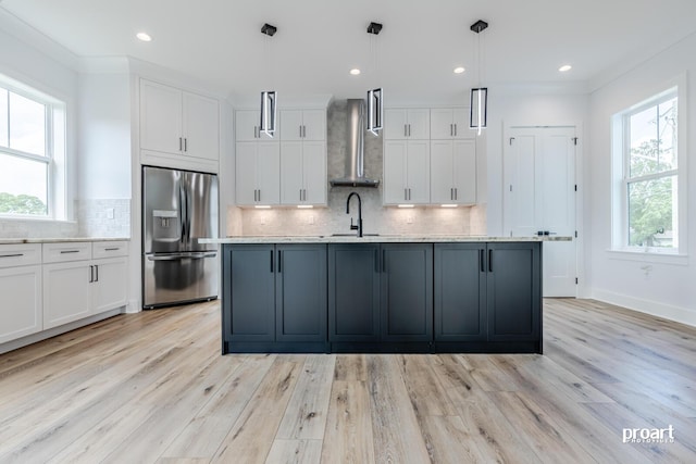 kitchen featuring white cabinetry, stainless steel fridge, hanging light fixtures, a center island with sink, and wall chimney exhaust hood