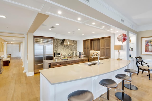 kitchen featuring sink, built in fridge, a large island with sink, wall chimney range hood, and a breakfast bar area