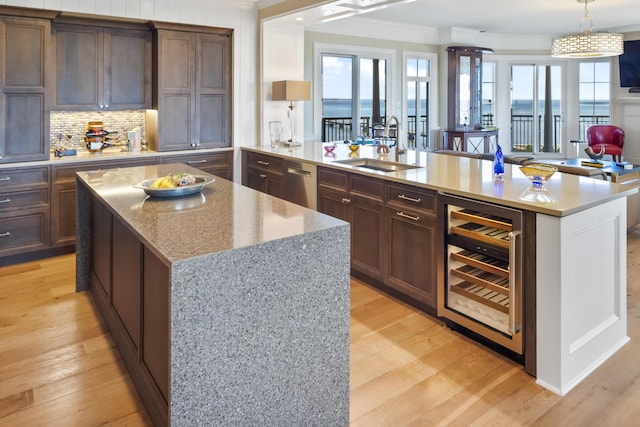 kitchen with light wood-type flooring, a kitchen island with sink, sink, light stone counters, and beverage cooler