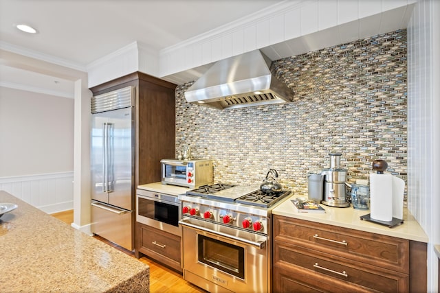 kitchen featuring backsplash, wall chimney range hood, premium appliances, light hardwood / wood-style floors, and crown molding