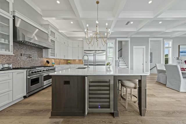 kitchen featuring sink, beverage cooler, premium appliances, wall chimney range hood, and a center island with sink