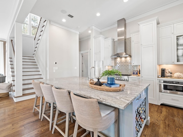 kitchen with stainless steel appliances, white cabinets, light stone countertops, wall chimney exhaust hood, and an island with sink