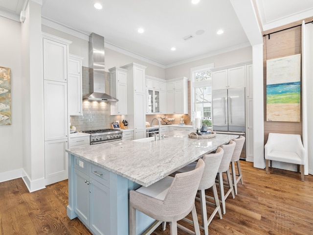 kitchen featuring a center island with sink, high quality appliances, wall chimney range hood, white cabinetry, and a sink