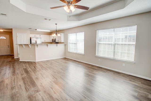 unfurnished living room featuring ceiling fan with notable chandelier, light hardwood / wood-style flooring, a raised ceiling, and a textured ceiling