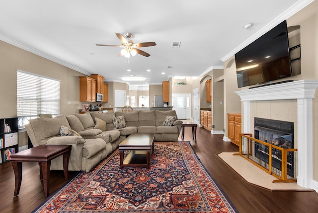 living area featuring dark wood-type flooring, ornamental molding, a tiled fireplace, and a ceiling fan