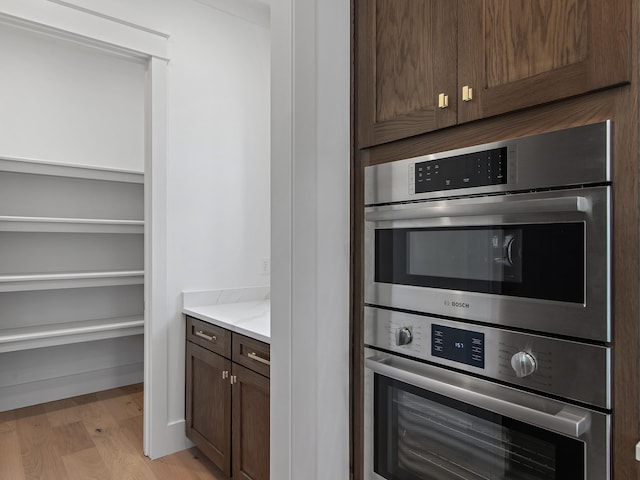 kitchen with dark brown cabinets, stainless steel double oven, light stone counters, and light hardwood / wood-style floors