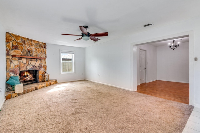 unfurnished living room with a stone fireplace, ceiling fan with notable chandelier, carpet flooring, visible vents, and baseboards