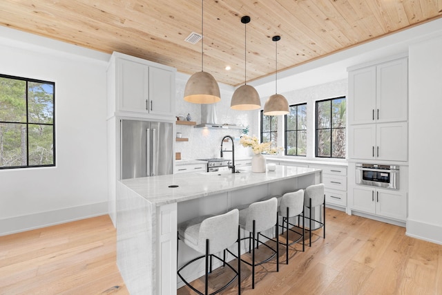 kitchen with appliances with stainless steel finishes, white cabinetry, wood ceiling, light stone countertops, and a center island with sink