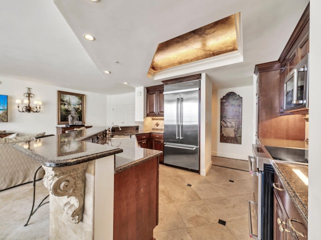 kitchen with a raised ceiling, appliances with stainless steel finishes, a kitchen breakfast bar, dark stone countertops, and recessed lighting