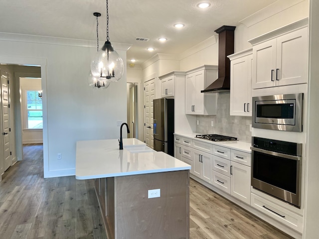 kitchen with an island with sink, wall chimney exhaust hood, stainless steel appliances, crown molding, and backsplash